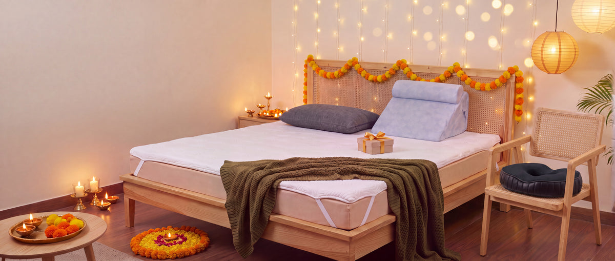 A bedroom dressed for the festive season with a marigold string on the headboard, fairy lights, diyas and a petal rangoli: a grey Midnight Serene memory-foam pillow, the light-blue TriAdjust wedge, the WhisperSleep topper and the Circle Ease donut cushion on a cane armchair